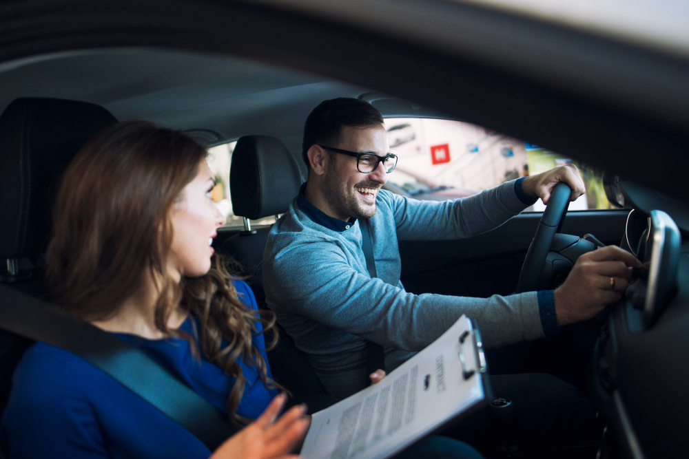 A smiling man drives a car while a woman sits in the passenger seat with a clipboard, both wearing seat belts during a test drive or car viewing.