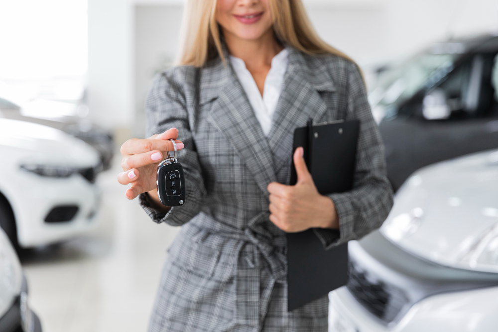 Salesperson in a plaid suit offers a car key on a showroom floor with other cars in the background and a clipboard in hand.] ,?