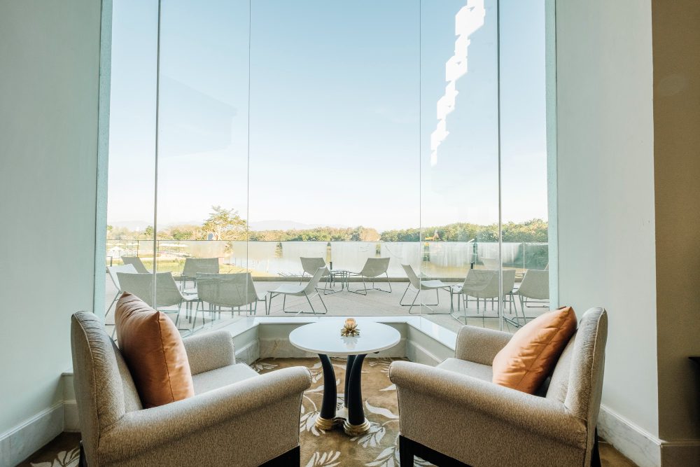 Cozy seating nook with two beige armchairs and orange cushions by a large glass window overlooking an outdoor patio and water.