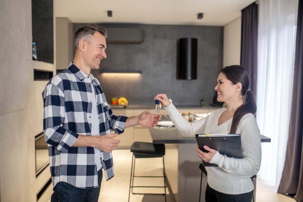 Woman hands keys to a man in a modern kitchen, both smiling during a home handover.