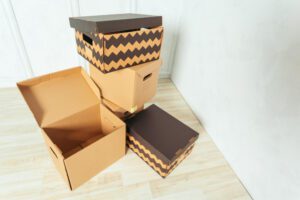 Pile of cardboard moving boxes stacked in a light room, some open or closed, on a wooden floor.