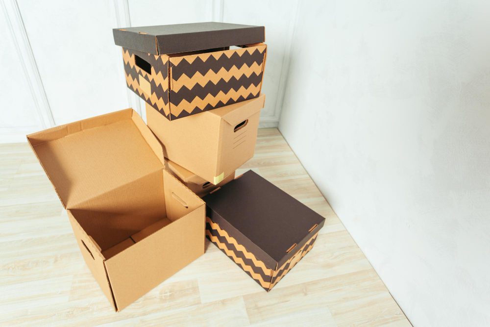 Pile of cardboard moving boxes stacked in a light room, some open or closed, on a wooden floor.