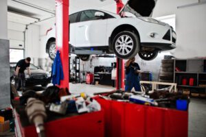 White car raised on a lift in an auto repair shop, with technicians and tools around far background in view.
